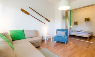 A modern living room with a beige sofa and a blue armchair. Oars hang on the wall, and in the background, a minimalist bedroom is visible.