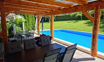 A cozy outdoor area with a wooden gazebo and a dining table. In the background, there is a blue swimming pool and well-kept greenery.