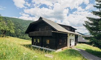 A charming wooden house in a green landscape, surrounded by gentle hills. The sky is clear with some clouds.