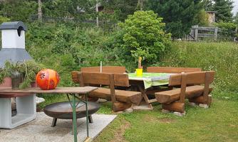 A rustic garden area with a wooden table and benches. In the background, there is a grill and a small tree.