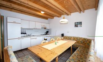 A bright kitchen with a large wooden table and benches. The walls have light wood details and there are modern kitchen appliances.