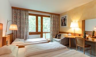 A cozy hotel room with two beds and large windows. The room is bright and inviting, featuring a small table and chairs as well as decorative elements.