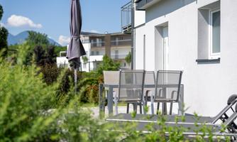 An elegant terrace with modern chairs and a sunshade. In the background, green trees and a blue sky can be seen.