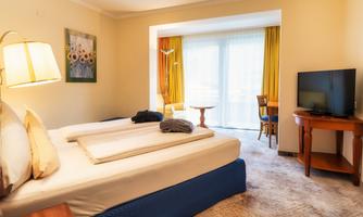 A cozy hotel room with two beds and a television. Large windows let in plenty of light, and there is a small table with chairs.