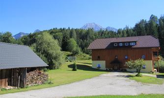 A rustic farmhouse with a red roof and a wooden barn. In the background, green meadows and tall mountains stretch under a clear blue sky.