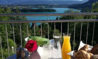 A beautiful breakfast table with cheese, cold cuts, and fresh rolls. In the background, one can see a picturesque lake and mountains under a clear blue sky.