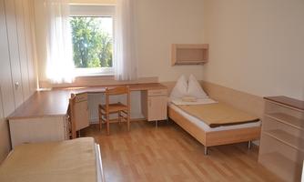 A simple room with two beds and a desk. Daylight shines in through the window.
