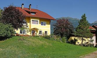 A charming yellow house with a red roof stands on a green hill. In the background are mountains and more trees.