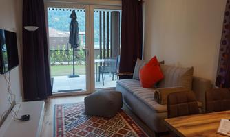 A cozy living room setup with a gray sofa and orange decorative cushions. The large window front offers a view of a balcony and the surrounding landscape.