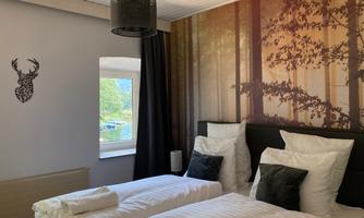 A cozy bedroom with two beds and a large window front. The wall is decorated with a tranquil forest image.