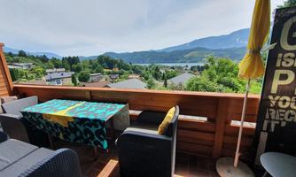 A cozy terrace with a table and chairs, overlooking mountains and a lake. The surroundings are green, and a sunshade is ready.
