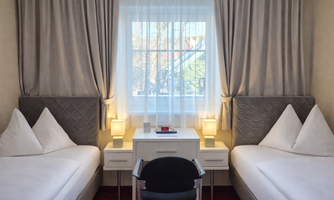 A cozy hotel room with two single beds and a desk. Large windows with curtains let in plenty of daylight.