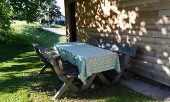 A wooden table with a blue tablecloth stands outdoors under a tree. Next to the table are several bench spots on a green meadow.