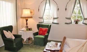 A cozy room with two green armchairs and a small table. The room has large windows with curtains and a warm, inviting atmosphere.
