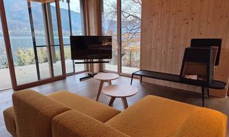 A modern living room with a yellow sofa and a television. Large windows offer a view of the nature outside.