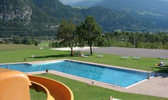 A swimming pool surrounded by green meadows and mountains. The sun is shining, and some loungers are ready for relaxation.