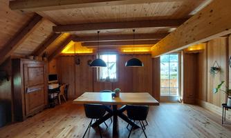 A cozy room with wooden walls and a large, bright window. Black chairs are around the table, and the ceiling is fitted with wooden beams.