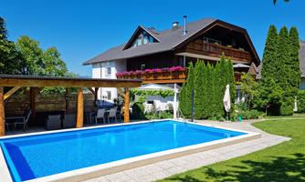 A beautiful swimming pool in a well-maintained garden with plenty of greenery. In the background, there is a modern house with balconies and flowers.