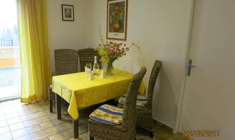 A cozy dining area with a yellow table and four chairs. On the table are flowers and bottles, while large windows let in plenty of daylight.