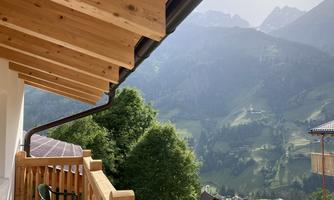 A cozy porch with wooden cladding and a view of the mountains. The landscape is green and hilly, surrounded by majestic peaks.