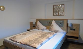 A modern bedroom with a large double bed and cozy wood paneling. The walls are painted in soft colors, and there is a wall decoration featuring a tree motif.