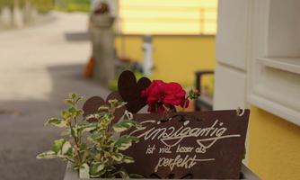 A flower pot with red flowers is in front of a yellow wall. In the background, a quiet path with houses and mountains can be seen.