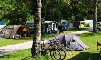 A campsite with various tents and trailers in a green environment. Some bicycles are on the lawn while vacationers enjoy nature.