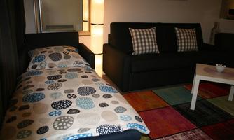A cozy living space with a sofa bed and a black sofa. The carpet has a colorful geometric pattern and a lamp provides pleasant light.