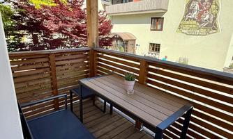 A beautiful balcony with a wooden table and several chairs. In the background, colorful trees and a building with a mural can be seen.