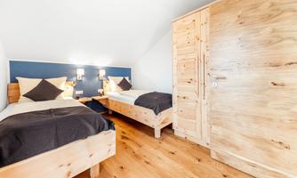 A cozy room with two wooden single beds and beautiful cushions. The walls are light blue, and there is a wooden wardrobe as well as warm light.