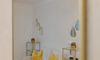 A modern living room with a comfortable white sofa and yellow cushions. There are golden shelves and stylish lighting.