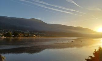 A tranquil landscape with a lake, surrounded by mountains and the warm evening sun. The sky is clear with fine streaks of clouds.