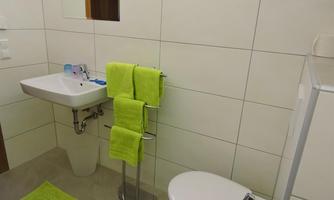 A modern bathroom with white tiles and bright green towels. It features a sink, a toilet, and a shower cabin.