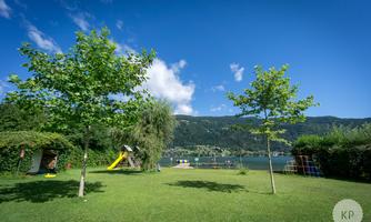A green meadow with two trees and a playground in the background. The lake and the mountains are visible in the distance, under a clear blue sky.