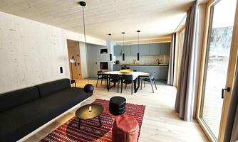 A modern living room with a black sofa and a rug in warm colors. The open kitchen is well-equipped and light streams in through large windows.