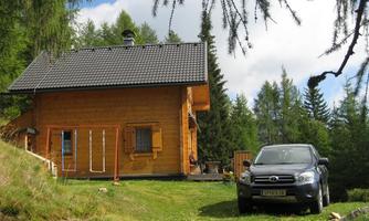 A cozy wooden house surrounded by trees in a green landscape. A black car is parked in front of the house.