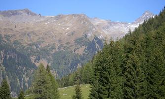 A picturesque mountain landscape with green forests and snow-capped peaks. The sky is clear and blue.