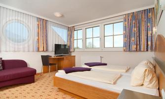 A bright hotel room with two single beds and a cozy sofa. Large windows provide plenty of daylight and offer a view outside.