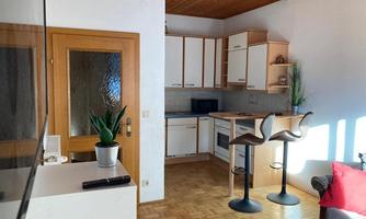 A small, modern kitchen with a dining area featuring two bar stools. The room has large windows that let in light and a cozy atmosphere.
