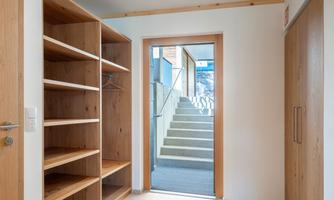 A bright hallway with an open wooden shelf and a door leading to a staircase. The walls are painted in neutral colors and the floor is tiled.