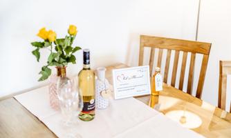 A beautifully laid table with a vase full of yellow roses, a bottle of wine, and a small sign. The atmosphere is inviting and cozy.