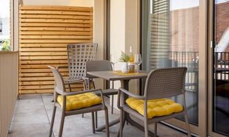 A cozy balcony with a table and four chairs. The chairs have yellow cushions and in the background, wooden paneling is visible.