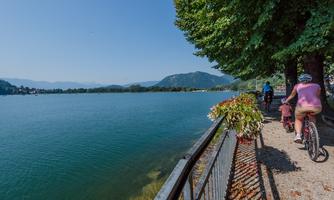 A picturesque lake landscape with cyclists on a path. In the background, there are mountains and a clear blue sky.