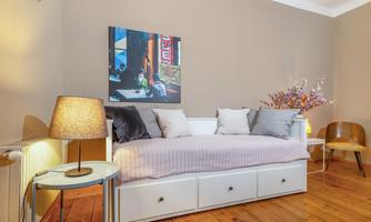 A cozy room with a daybed and decorative pillows. A colorful painting hangs on the wall, and a table with a lamp provides warm light.