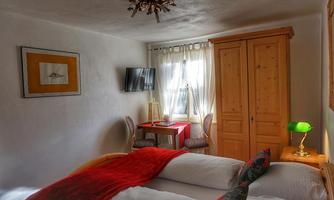 A cozy bedroom with two beds and red accents. There is a table, a chair, and a TV, as well as a wooden wardrobe in the room.