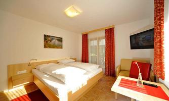 A cozy room with a double bed and a television. There is a comfortable armchair and large windows with red curtains.