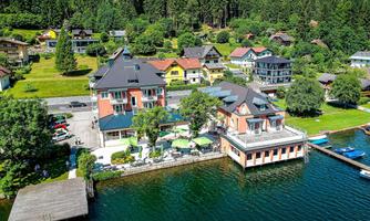 A picturesque building by the shore of a clear body of water, surrounded by green trees and hills. Several residential houses are located in the background, creating a tranquil, rural setting.