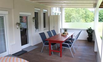 A cozy porch with a large red table and several chairs. In the background, you can see green trees and a tranquil atmosphere.