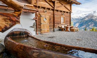 A cozy wooden cabin in the mountains with a flowing stream. In the background, the majestic mountains are visible.