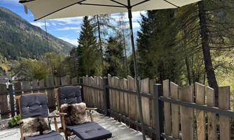 A cozy terrace with two lounge chairs and a sun umbrella. In the background, green mountains and a blue sky can be seen.